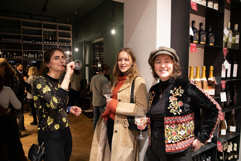 Drei junge Frauen mit Weingläsern in der Hand genießen einen Abend in einer modernen Vinothek, umgeben von Regalen voller Weinflaschen und stimmungsvoller Beleuchtung.