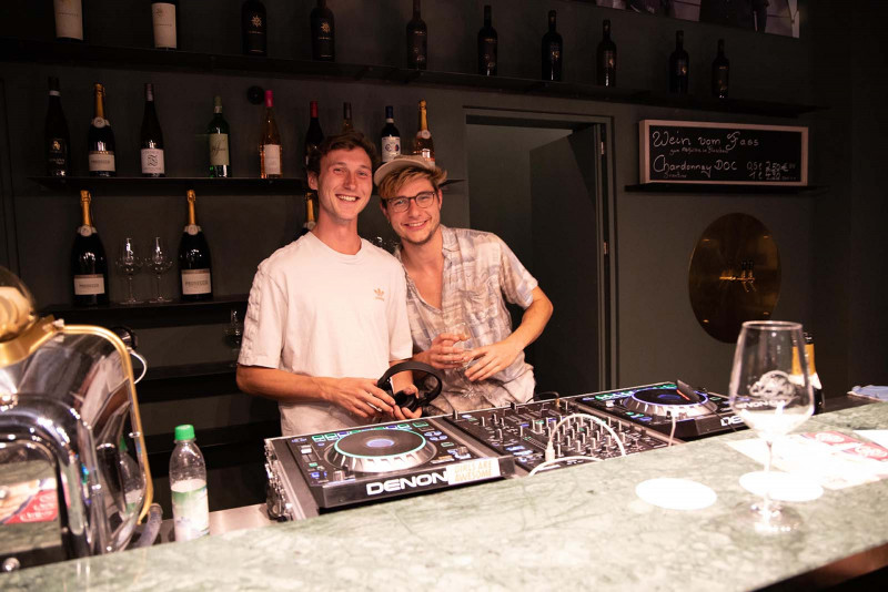 Zwei junge DJs bei der Eventreihe ‚Bubbles & Beats‘ in Saffer’s WinzerWelt, lachend hinter dem DJ-Pult in der Vinothek, mit Prosecco-Flaschen im Hintergrund und einem Weinglas auf der Theke.