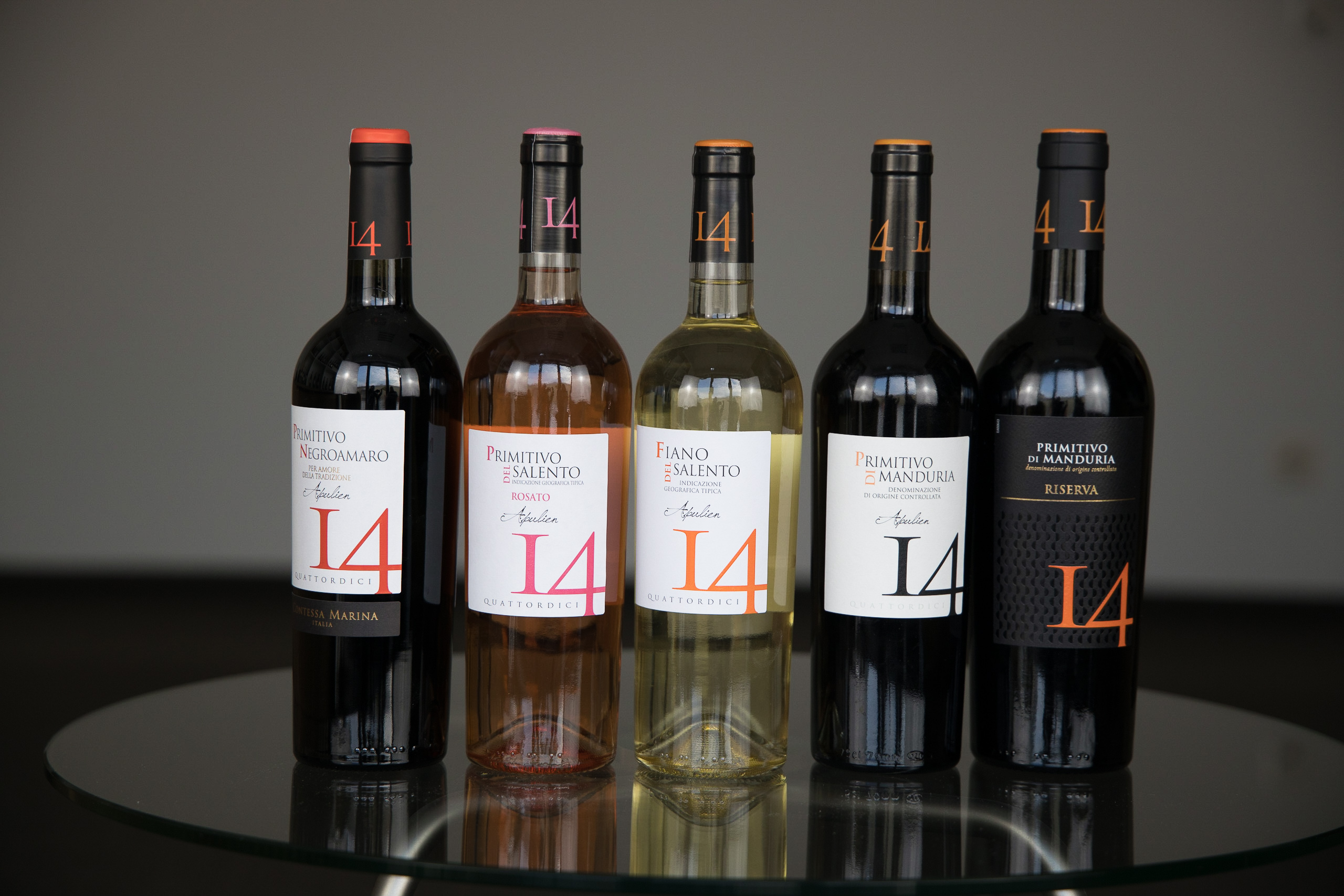 Fünf Weinflaschen der Linie 'I 4' auf einem Glastisch vor neutralem Hintergrund – Rotwein, Rosé und Weißwein mit modernem Etikettendesign in Weiß, Schwarz und Orange.