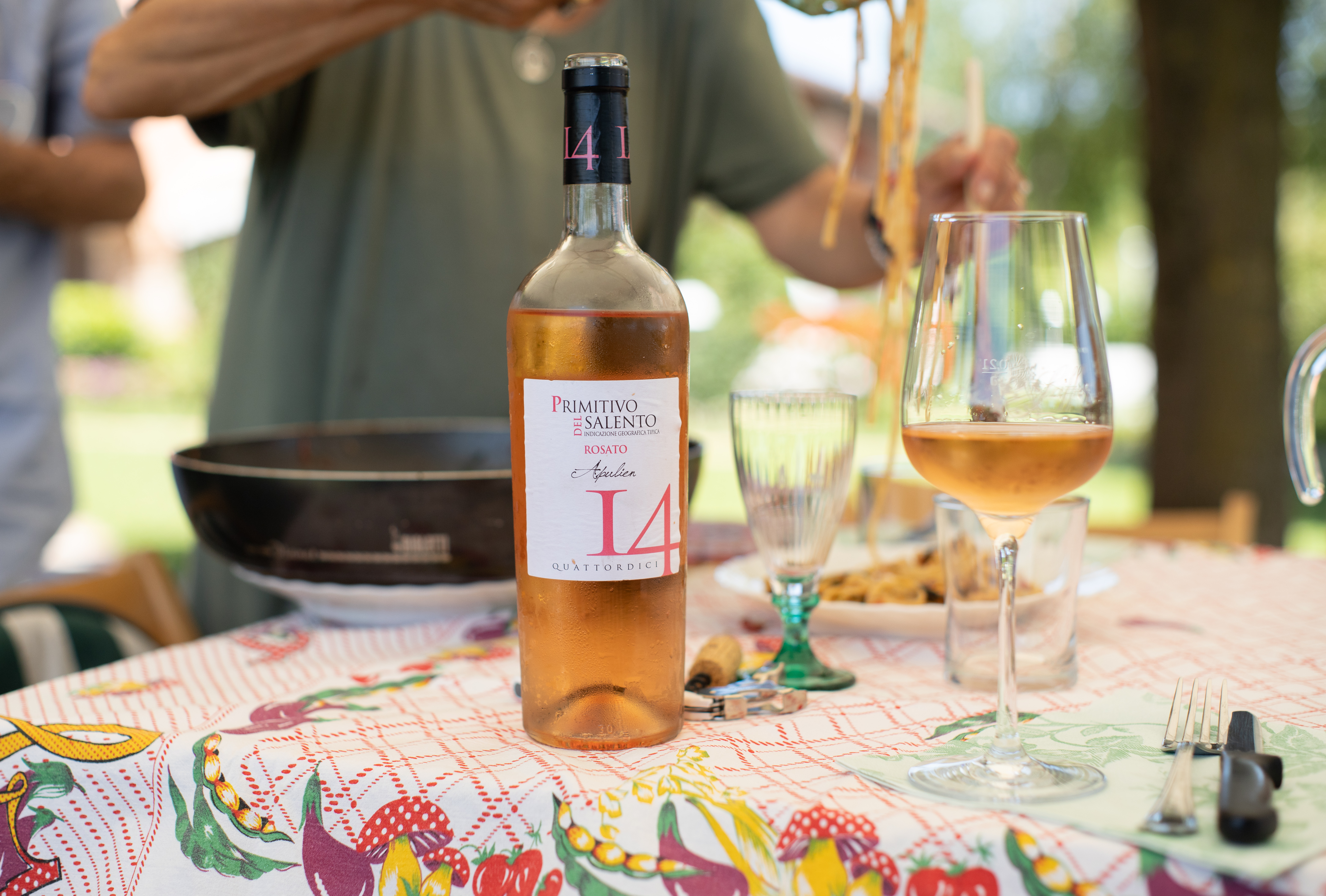 Flasche Roséwein 'I 4 Primitivo Rosato' auf einem sommerlich gedeckten Tisch mit bunchem Tischtuch, Weinglas und Pasta – im Hintergrund eine Person beim Servieren im Freien.